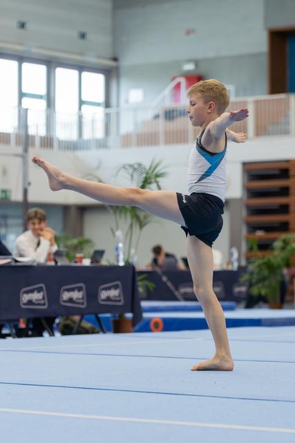 Young boy performs a high leg raise on the floor exercise mat, arms extended, focused and balanced at a gymnastics meet.