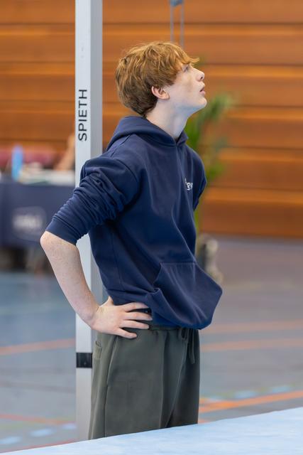 Red-haired teen gymnast looks upward with hands on hips, focused and composed beside a Spieth apparatus in a gym.