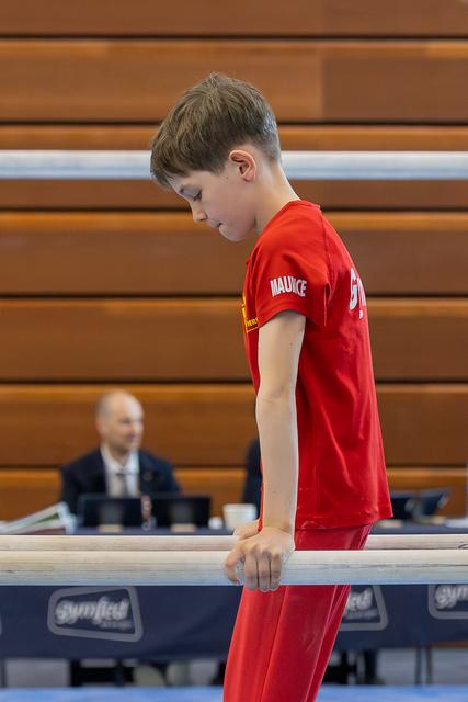 Boy named Maurice in red looks down with focus, gripping parallel bars at a gymnastics competition.