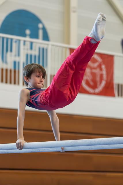 Focused young boy performs a straddle hold on parallel bars, legs raised high, showing strength and concentration at an indoor gymnastics meet.