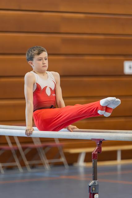 Young boy in red leotard performs a straddle hold on parallel bars, legs extended, expression focused and determined.