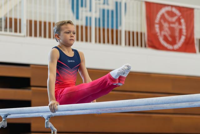 Focused young gymnast performs a leg raise on parallel bars, wearing a navy and red Quatro leotard in a gymnasium.
