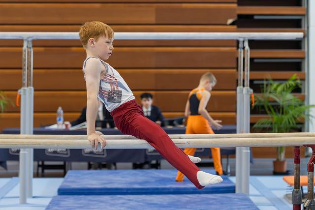 Red-haired boy sits intently on parallel bars, legs extended, focused expression during a gymnastics meet.