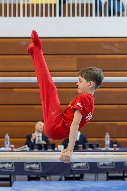 Young boy in red performs a straddle hold on parallel bars, face calm and focused during a gymnastics meet.