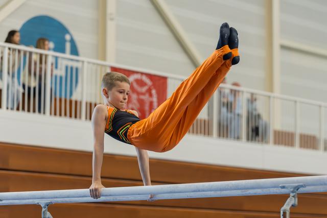 Young boy in orange performs a precise leg raise on parallel bars, focused expression, indoor gymnastics hall.