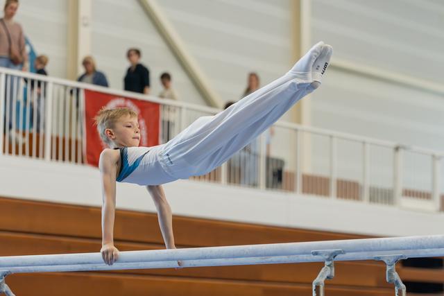 Focused young boy performs a pike hold on parallel bars, legs raised high, showcasing strength and concentration at a gymnastics meet.