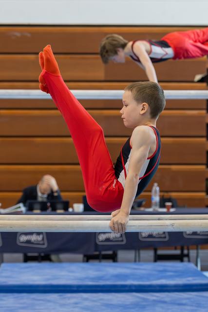 Young boy in red gymnastic suit holds a pike position on parallel bars, displaying intense focus and strength.