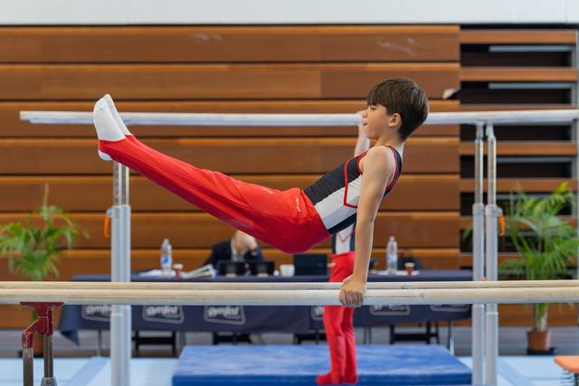 Focused young male gymnast holds a straddle L-sit on parallel bars, legs extended, expression intent during a meet.