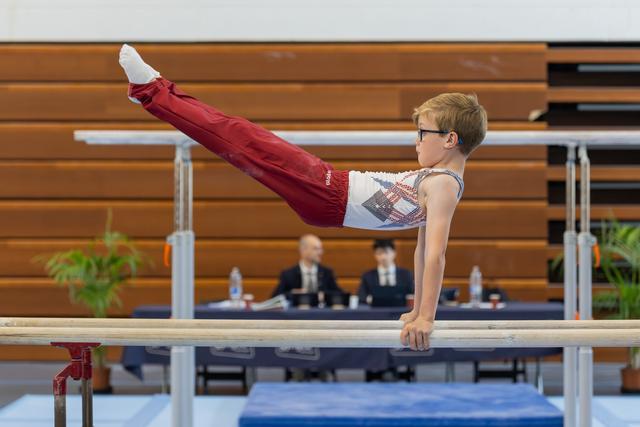 Young boy with glasses performs a straddle hold on parallel bars, legs raised high, expression intense and focused.