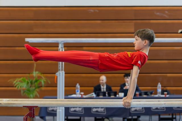 Focused young boy in red holds a perfect straddle-L on parallel bars before judges at a gymnastics meet.