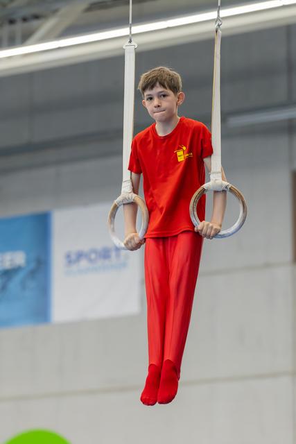 Young boy in red gymnastics suit holds still rings, expression calm and concentrated in an indoor sports hall.