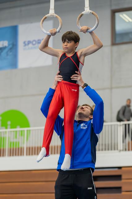 Young male gymnast grips still rings with focused expression while coach in blue jacket steadies him at the waist.