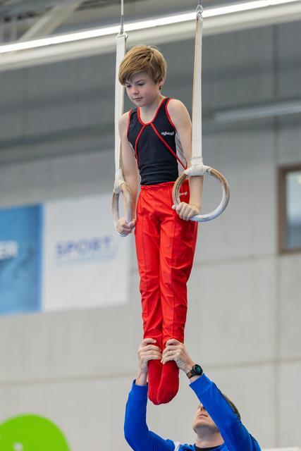Young boy performs on gymnastics rings with intense focus while a coach in blue supports his feet below.