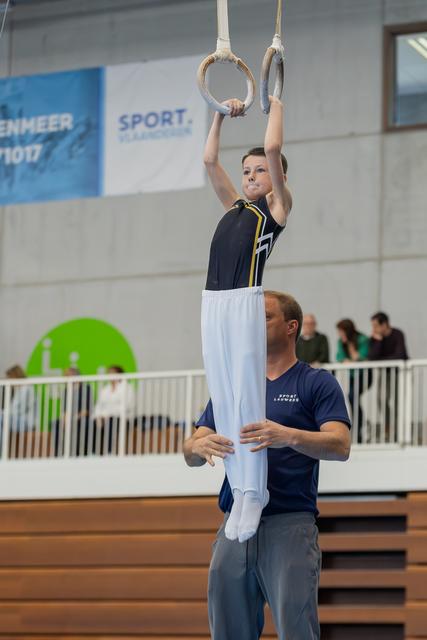Young boy grips gymnastic rings with focus while a coach steadies him from below in an indoor sports hall.
