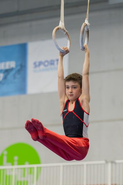 Young male gymnast hangs from still rings in a pike position, wearing red and black, with intense focus on his face.