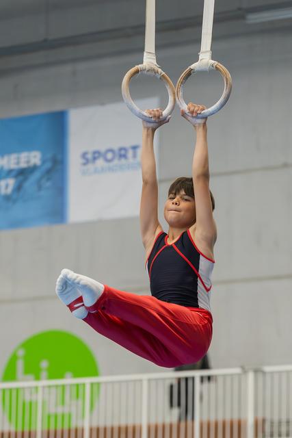 Young male gymnast hangs on still rings in a tucked pike position, focused expression, wearing red and black leotard.