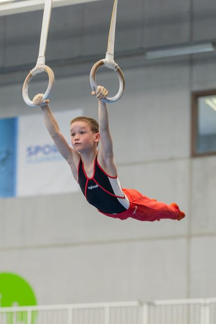Determined young boy grips gymnastic rings mid-air, body arched, wearing navy and red leotard in indoor sports hall.