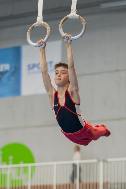 Young male gymnast hangs focused on still rings, body arched, wearing navy and red leotard in an indoor gym.