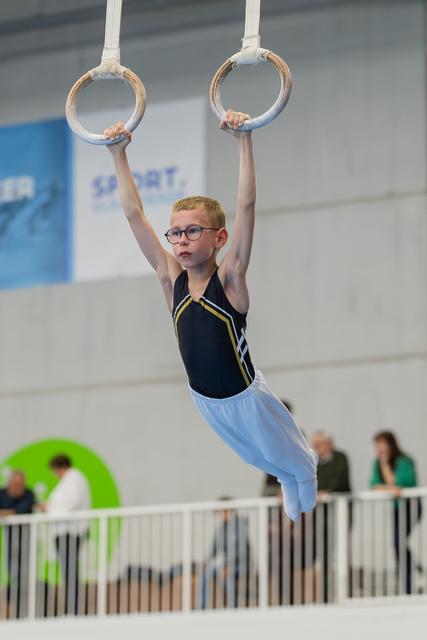 Young boy with glasses hangs with determination on gymnastic rings, body straight, in a sports hall during competition.