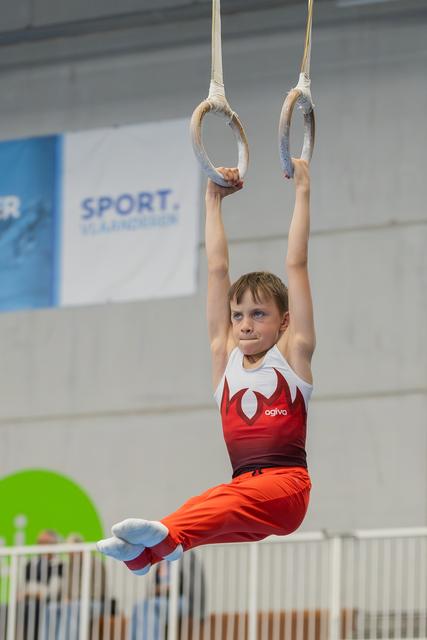 Young male gymnast hangs from still rings in a tuck position, focused expression, wearing red and white agiva leotard.