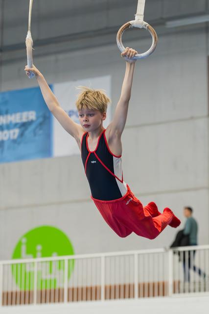 Blond boy hangs from gymnastic rings, legs tucked, focused expression, competing in an indoor gymnastics hall.