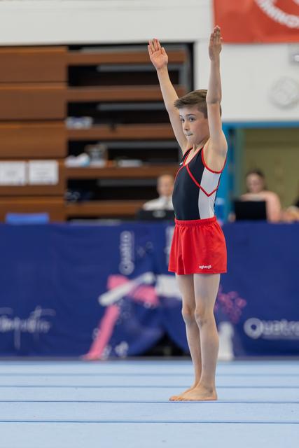Young male gymnast raises both arms in a confident salute on the floor exercise mat during a gymnastics meet.