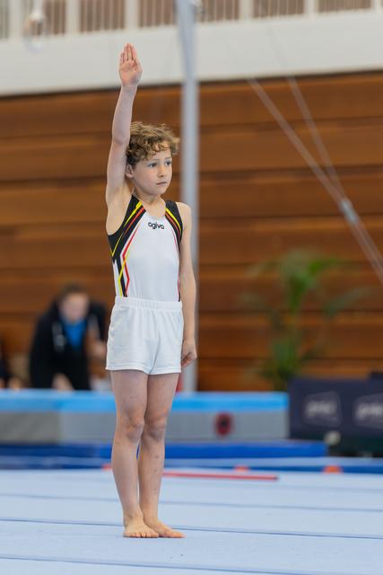 Young male gymnast raises hand to salute the judge, standing barefoot on the blue floor exercise mat, focused and composed.