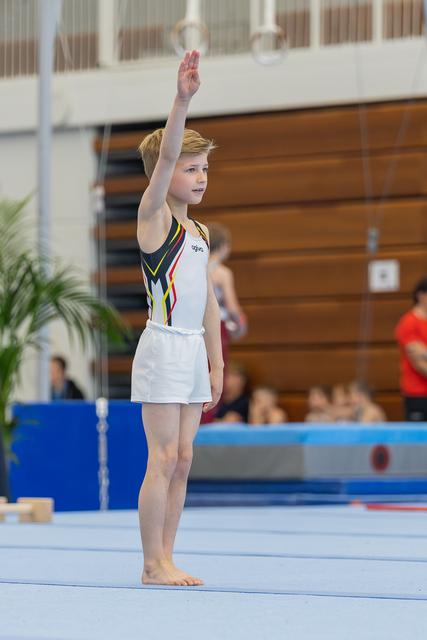 Young gymnast raises hand to salute judges before floor routine, standing barefoot on blue mat in gymnastics hall.