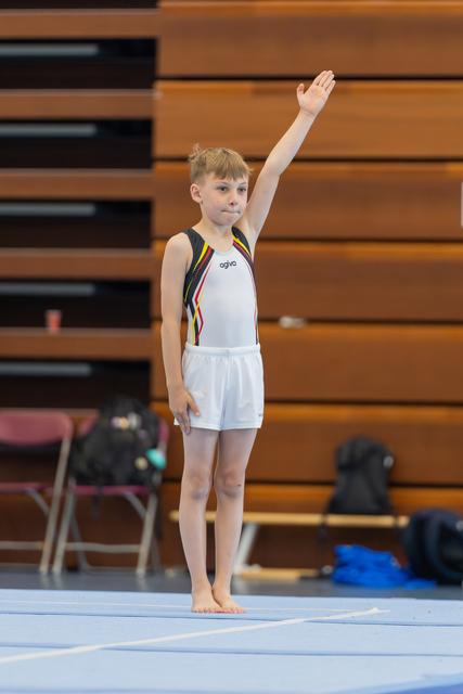 Young gymnast raises hand to salute judges, standing confidently barefoot on the floor exercise mat in a gym.