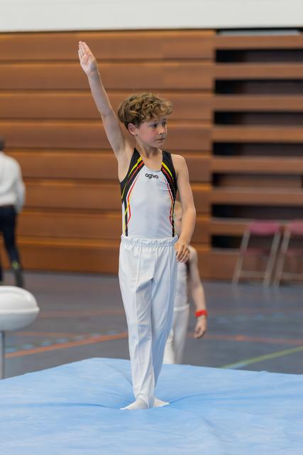 Young male gymnast raises one arm to salute the judges, standing barefoot on the floor exercise mat, composed and focused.