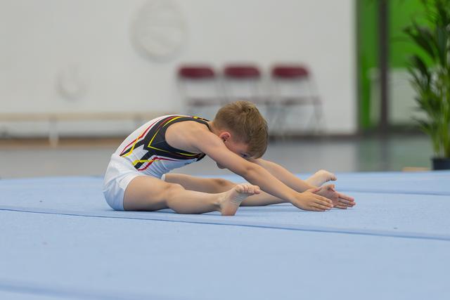 Young boy gymnast in black leotard performs a seated forward stretch on blue floor mat, focused and flexible.