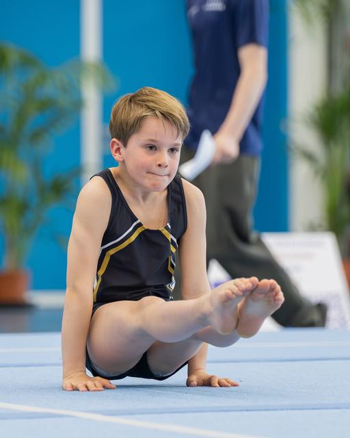 Young boy gymnast seated on blue mat, legs raised, looking focused and composed during a gymnastics event.