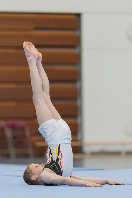 Young gymnast performs a shoulder stand on the floor mat, legs extended upward, during an indoor gymnastics event.