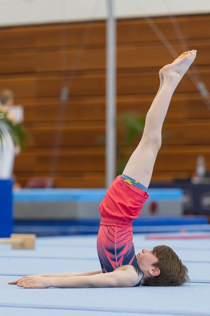 Young male gymnast performs a shoulder stand with one leg extended upward on a blue floor mat in a gymnasium.