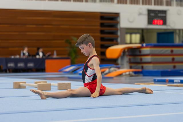Young boy gymnast performs a perfect split on the blue floor mat, looking down with intense concentration during a meet.