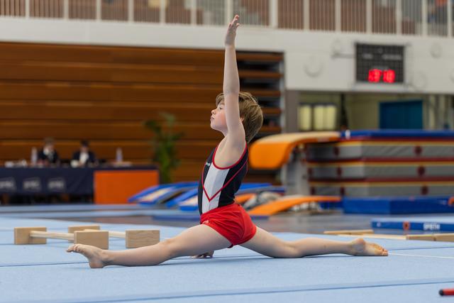 Young gymnast performs a split on the floor exercise mat, arm raised gracefully, expression calm and focused.