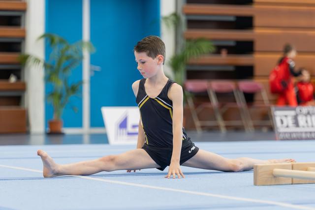 Young boy gymnast performs a perfect split on blue floor mat, focused expression, hands pressed to ground.