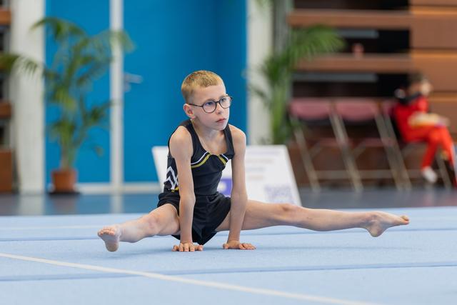 Young boy with glasses performs splits on blue gymnastics mat, focused expression, wearing black leotard during competition.