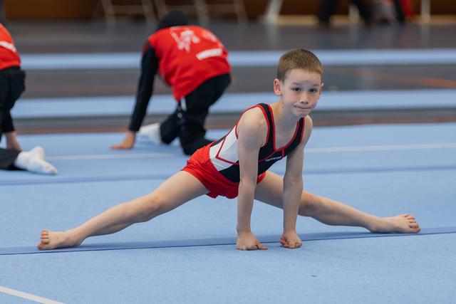 Young boy in red gymnastic suit performs a splits stretch on blue mat, looking focused and confident.