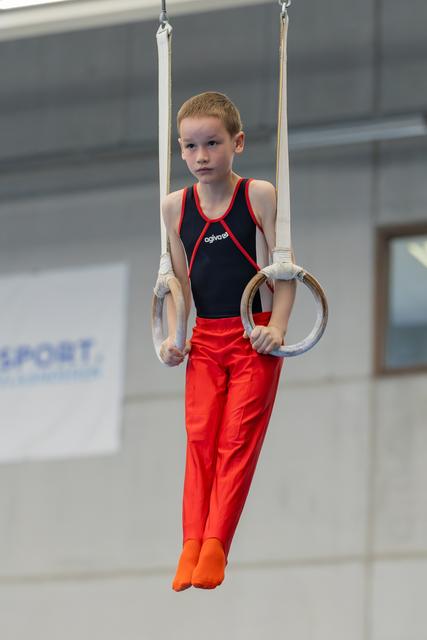 Young boy holds gymnastic rings with steady grip and focused expression, wearing red and black leotard in sports hall.