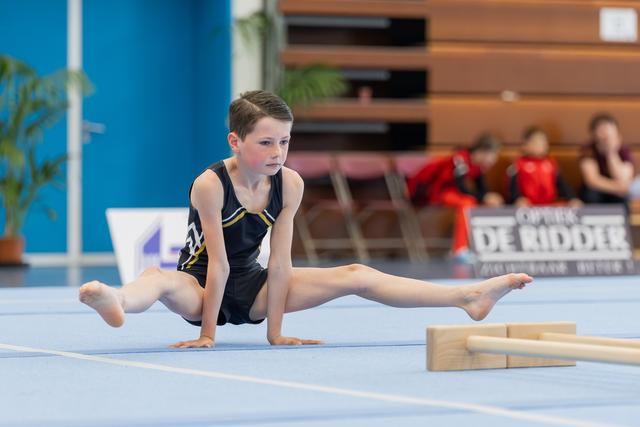 Focused young boy holds a straddle support position on the blue floor mat during a gymnastics competition.