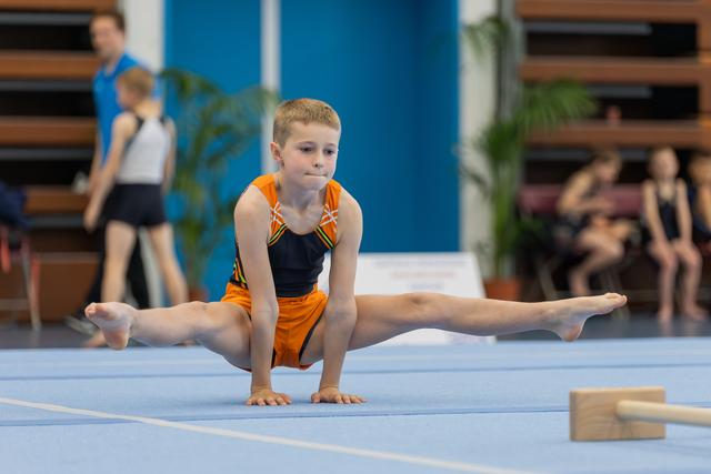 Focused young boy in orange leotard performs a straddle split on blue floor mat during gymnastics competition.