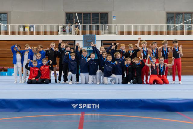 Young male gymnasts celebrate with raised fists and medals, posing joyfully in teams inside a gymnastics hall.
