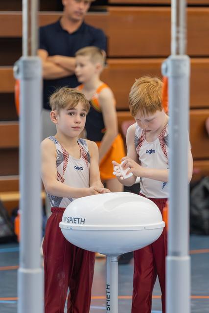 Two focused young gymnasts apply chalk at a Spieth apparatus, one helping the other, with teammates watching from bleachers behind.