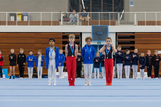 Four young gymnasts stand proudly on the floor mat, displaying their medals at an indoor gymnastics award ceremony.