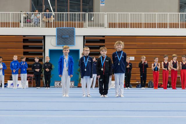 Four young gymnasts stand proudly on the gym floor wearing medals, surrounded by teammates clapping in the background.