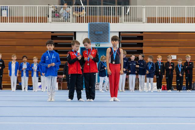Young male gymnasts stand on the floor mat during a medal ceremony, examining their medals, in a sports hall.