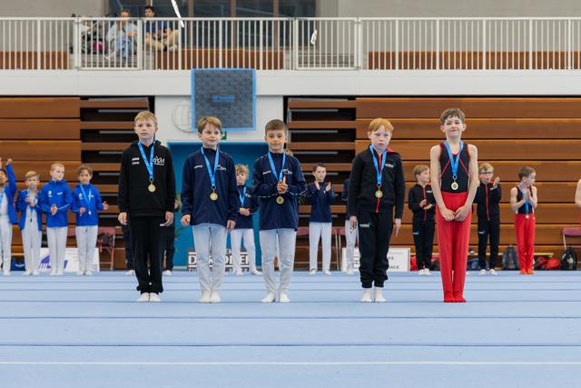 Five young male gymnasts stand proudly on the floor wearing gold medals, lined up at an indoor gymnastics competition.