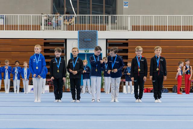Young male gymnasts stand proudly on a blue podium wearing gold medals at an indoor gymnastics competition.