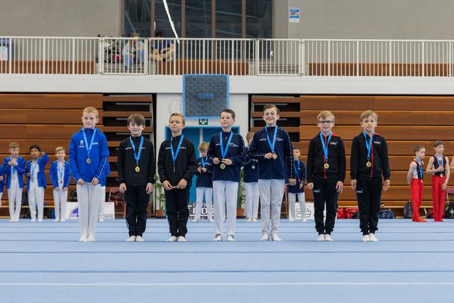 Seven young gymnasts stand proudly on the floor displaying their medals, smiling after a gymnastics competition.